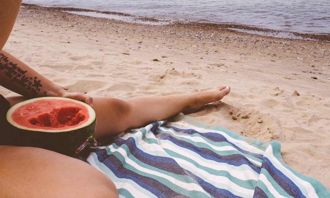 alimenti ricchi d'acqua | particolare di donna seduta in spiaggia su un asciugamano da mare mentre mangia metà anguria